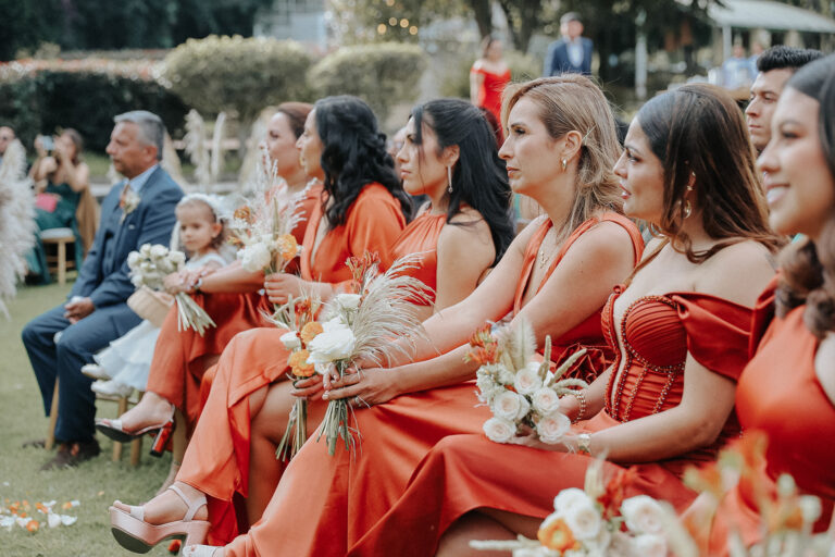 Invitadas a una boda con sus vestidos en color terracota con sus peinados y mirando al altar.